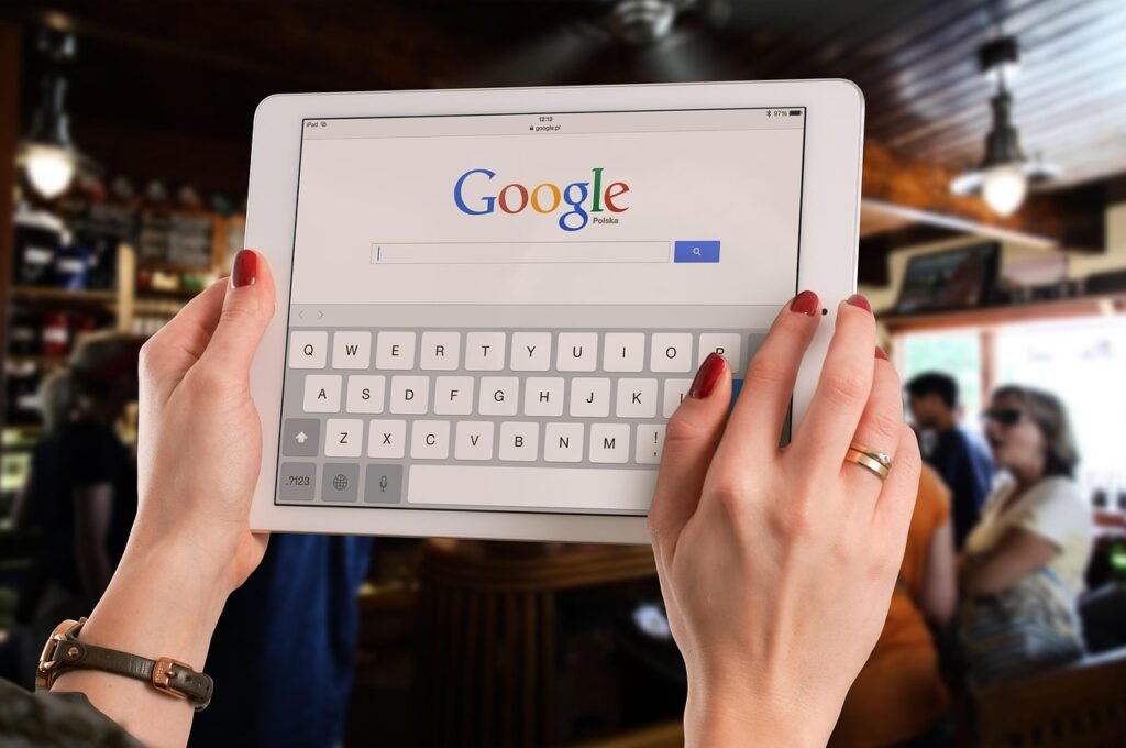 A person holds a white tablet featuring the Google Search homepage, set against a softly blurred café or restaurant background.