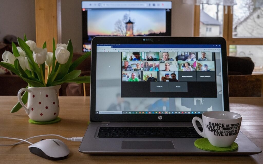 A laptop on a wooden table displays a Zoom meeting with multiple participants. Nearby are a vase of white tulips, a mouse, and a coffee mug. A TV screen is in the background.